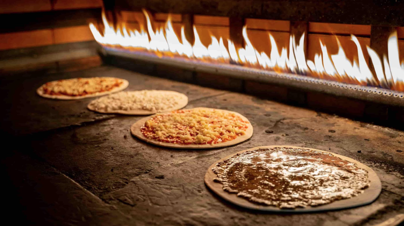 Four types of pizza being cooked by a fire.