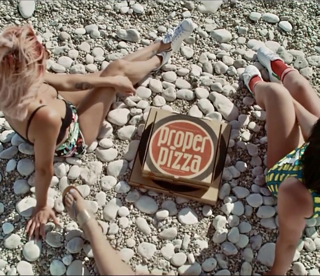 Two women on a stoney beach wearing swimsuits with two Proper Pizza boxes between them