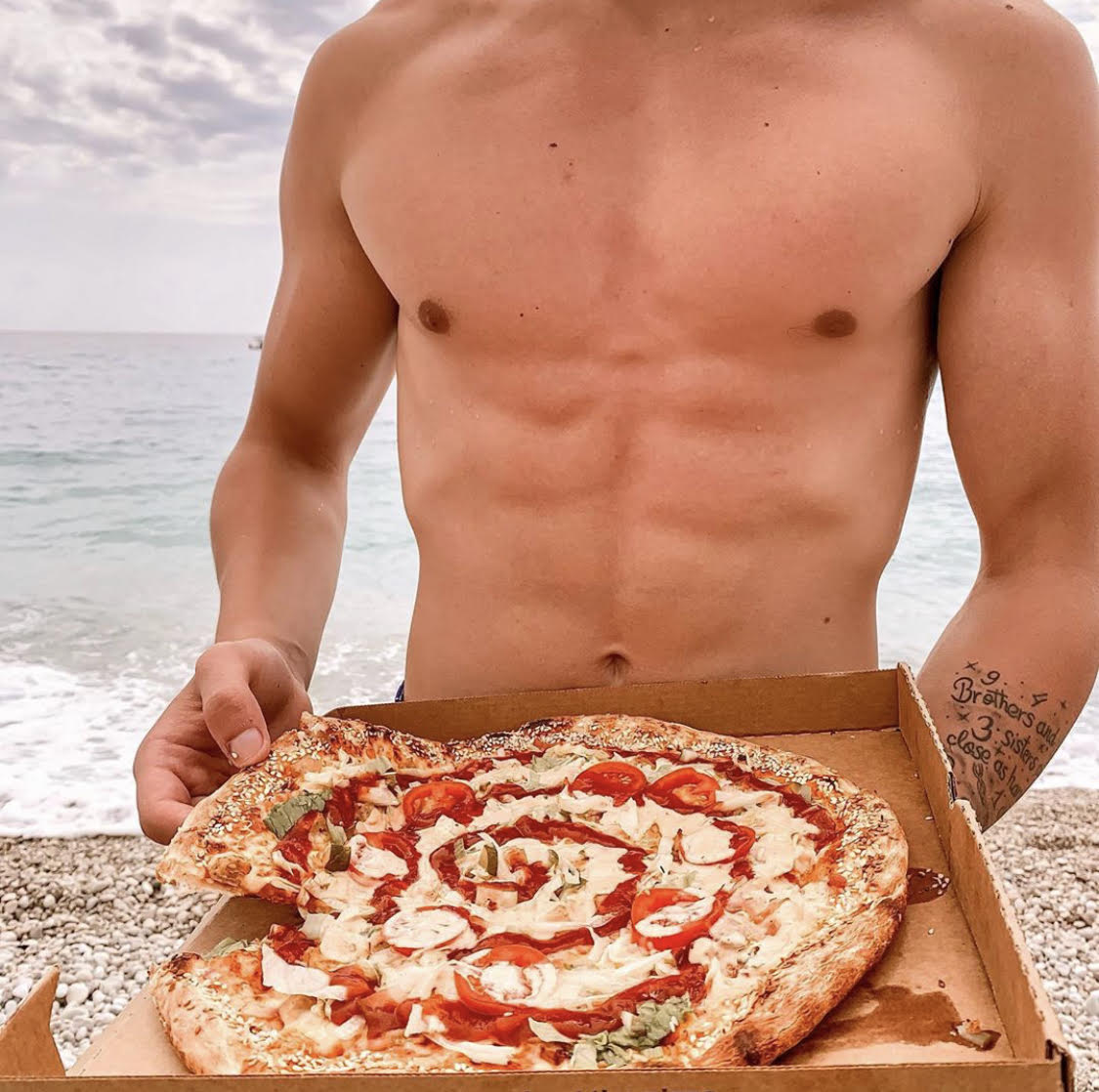 Shirtless man holding a slice of pizza on the beach